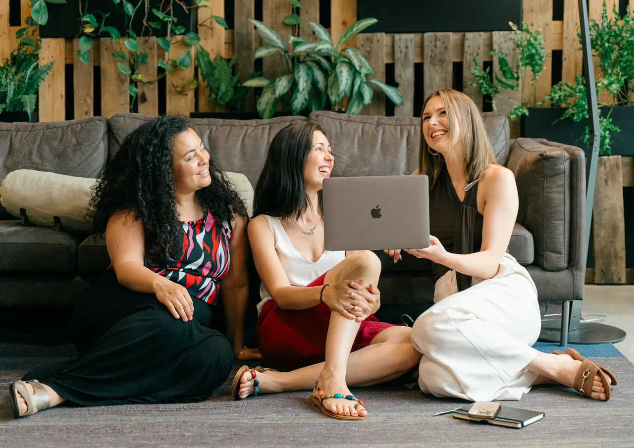 Three members laughing together on a sofa, around a laptop